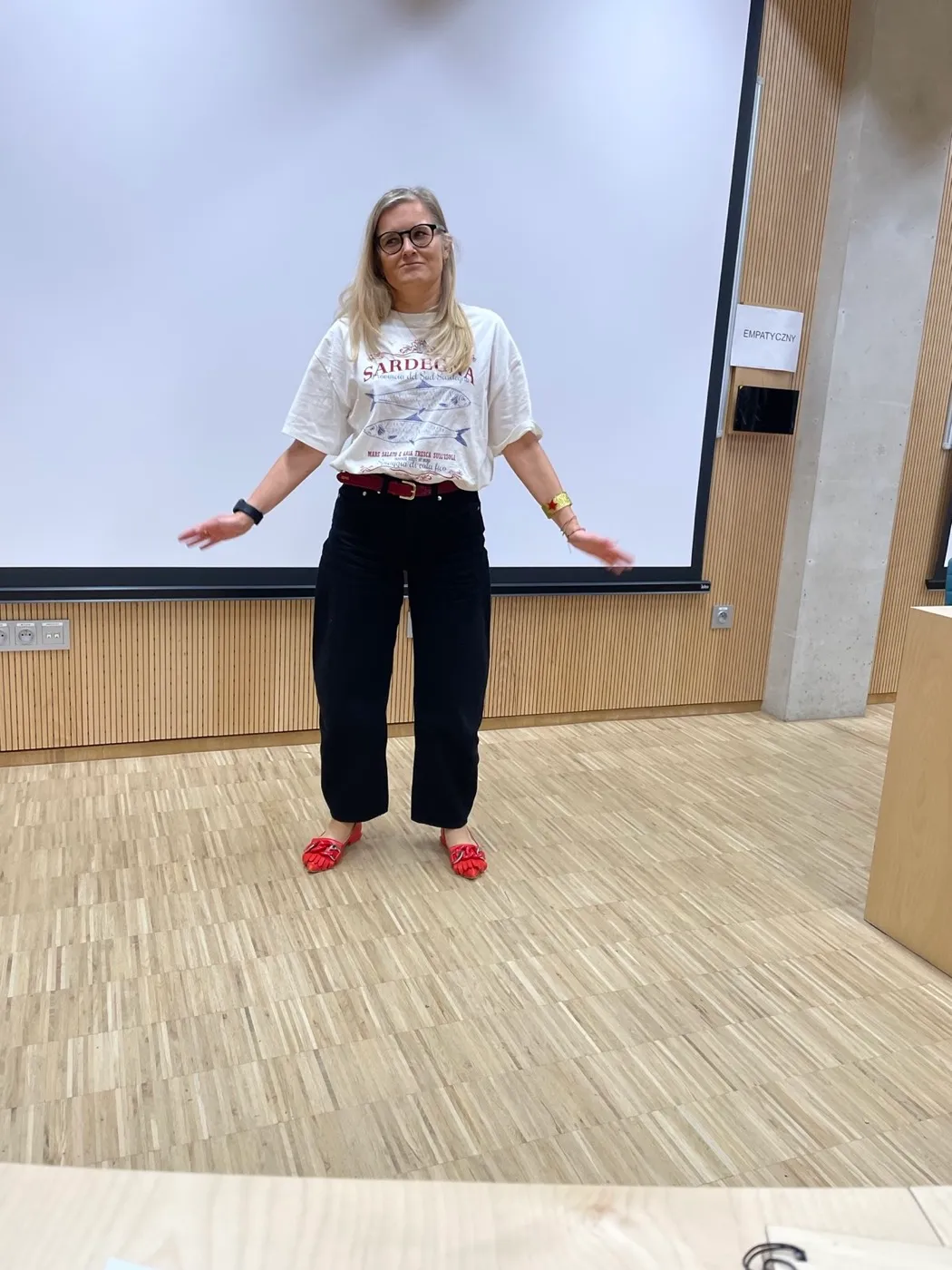 Mówczyni w czerwonym swetrze gestykuluje przed salą